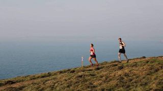 Two runners run down a grassy slope, the sea in the background.