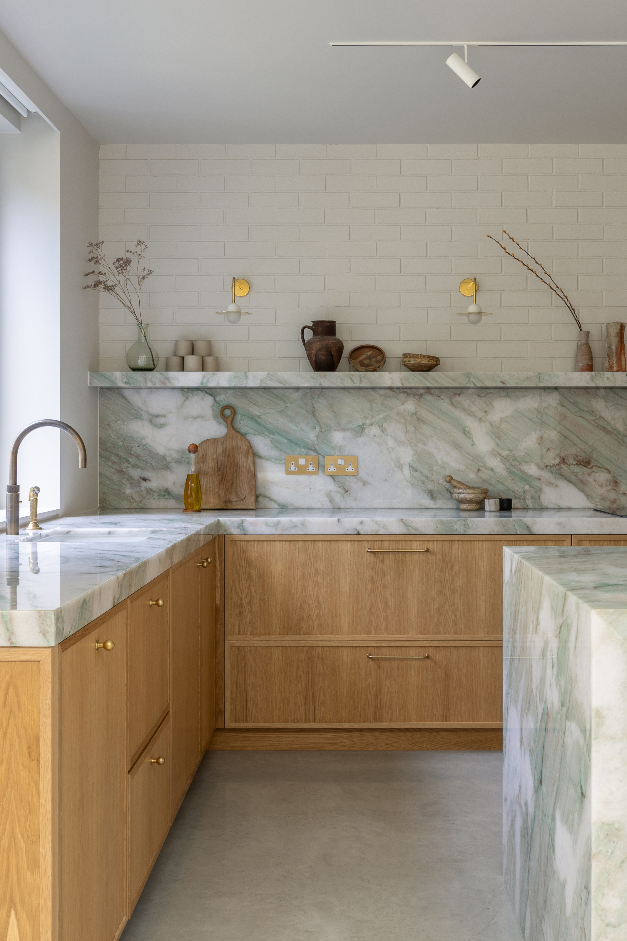 Kitchen with natural wood cabinets, white and gray-veined splashback and countertop, open shelving and painted brick wall