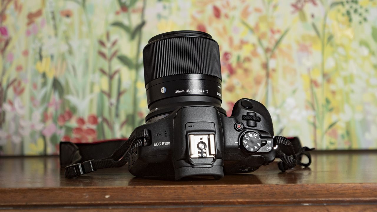 Sigma 30mm F1.4 DC DN Contemporary lens attached to a Canon EOS R100, on a wooden surface with floral backrop