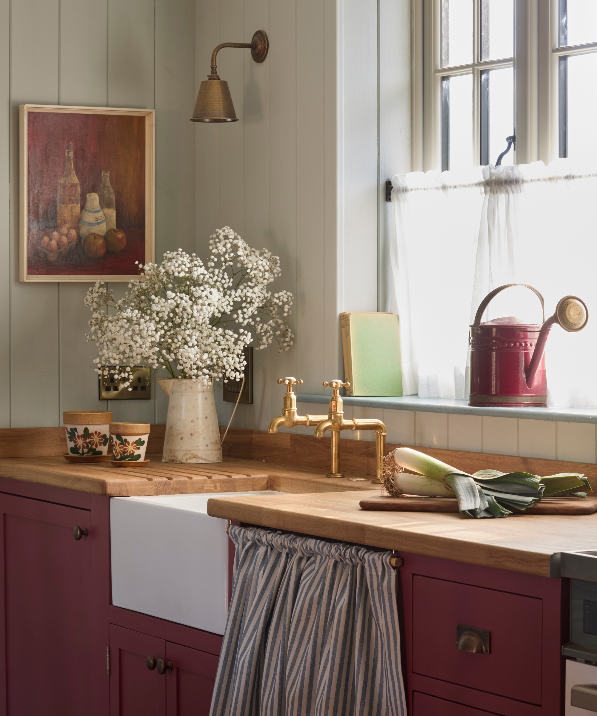 The corner of a costal cottage kitchen with light blue paneled walls, wood countertops, burgundy cabinets, and antique decor