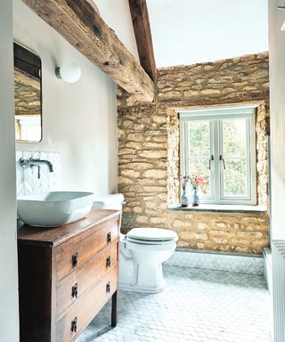 bathroom with exposed stone wall, wooden beams, tiled floors, toilet and sink on wooden vanity unit