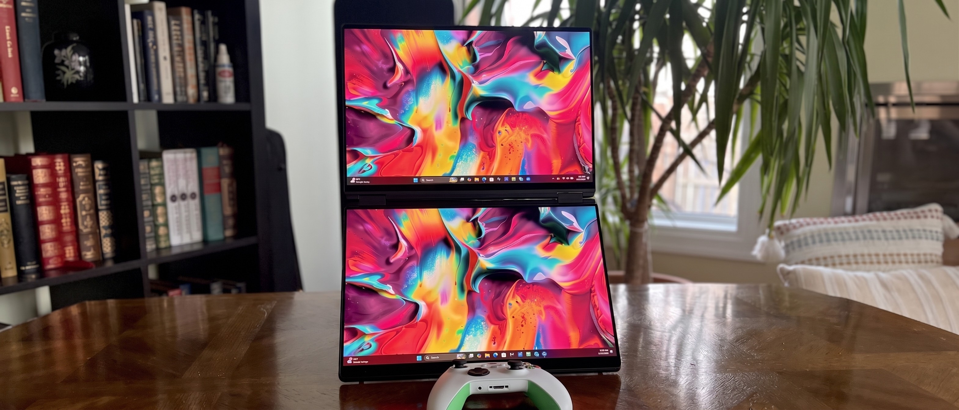 Dual monitors displaying vibrant abstract art with colorful swirls, placed vertically on a wooden table. A gaming controller is between them, with books and a plant in the background.