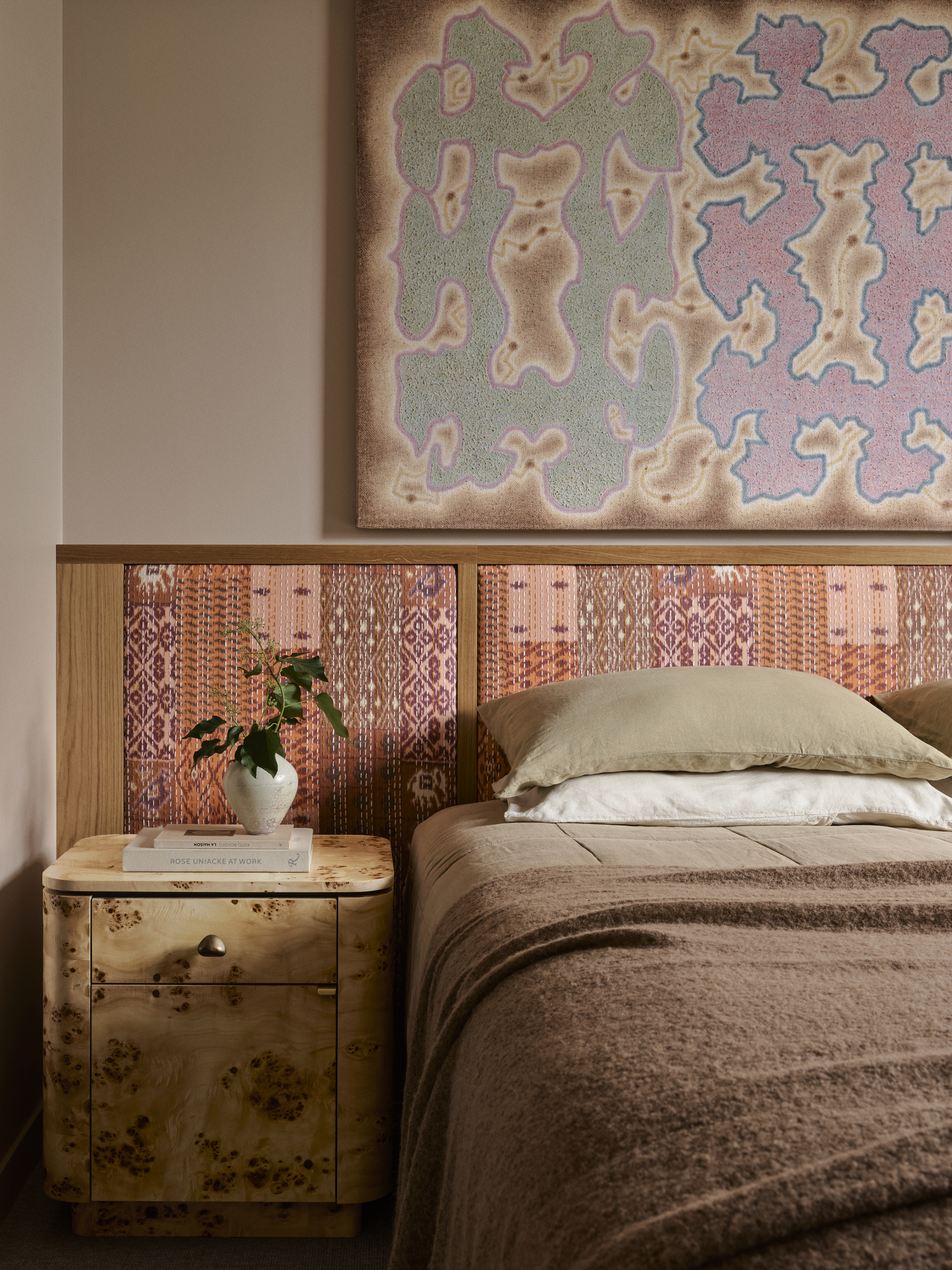 modern bedroom with plaster walls, large artwork, upholstered extended headboard with fabric, burl side table, bed made with brown bedding