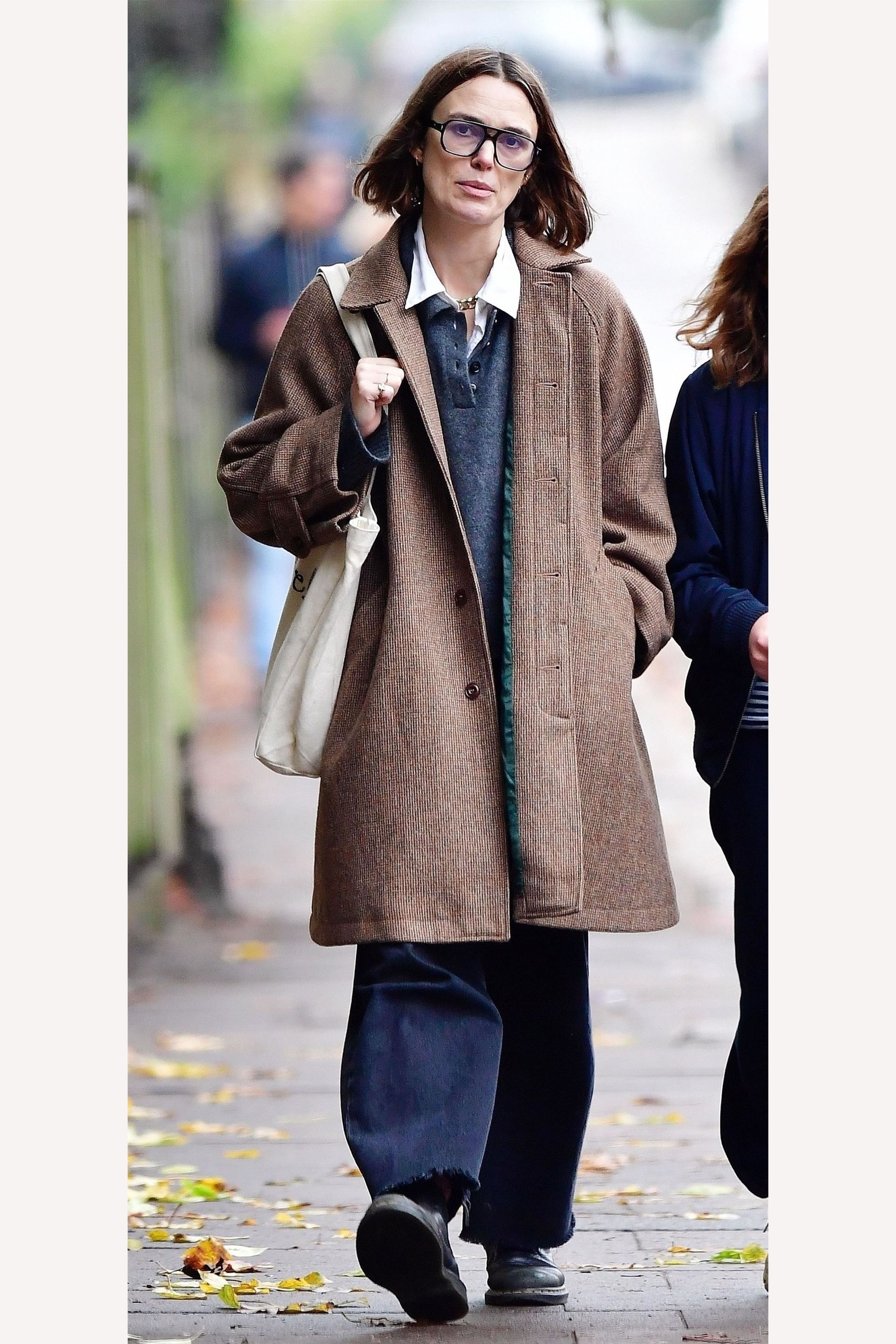 Keira Knightley wearing a brown coat, navy polo, dark-wash jeans, and black boots.