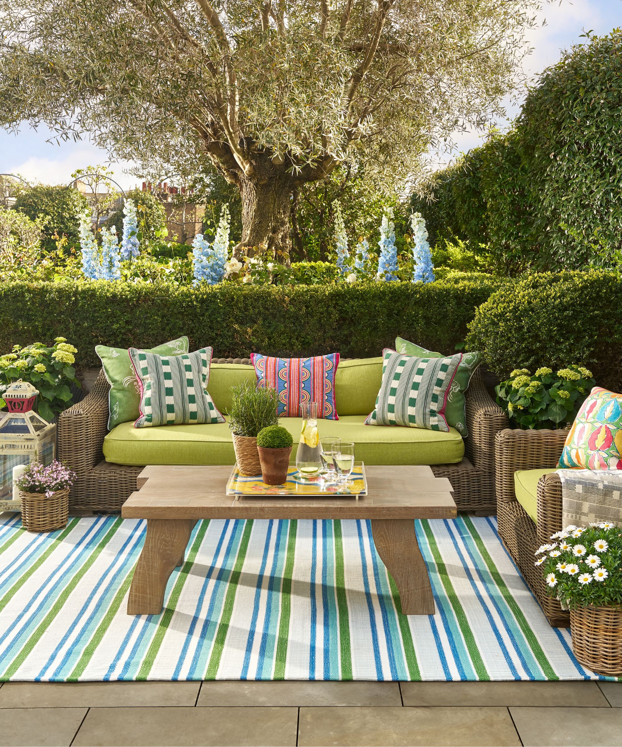 outdoor living room area with a blue and green striped rug, wooden coffee table, wicker sofas with green cushions and patterned throw pillows