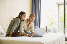 Medium wide shot of smiling couple relaxing on bed using laptop to plan vacation travel