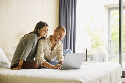 Medium wide shot of smiling couple relaxing on bed using laptop to plan vacation travel