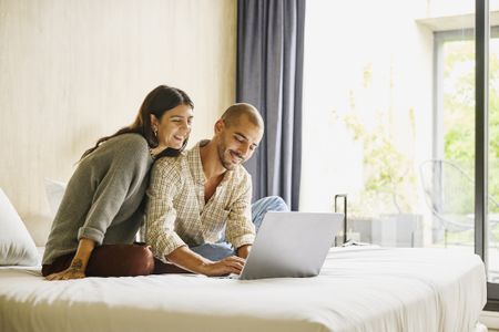 Medium wide shot of smiling couple relaxing on bed using laptop to plan vacation travel