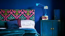 A bedroom painted deep midnight blue with double bed, patterned fabric bedhead and base, and rich jewel coloured bedding and furniture.