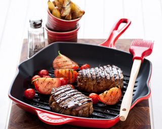 A red Le Creuset griddle pan with steak and grilled tomatoes, with silicone brush and ramekins with potato wedges
