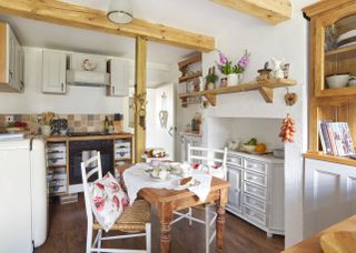 a small freestanding kitchen, with mostly white units, wooden beams on the ceiling, a small dining table and a small white fridge