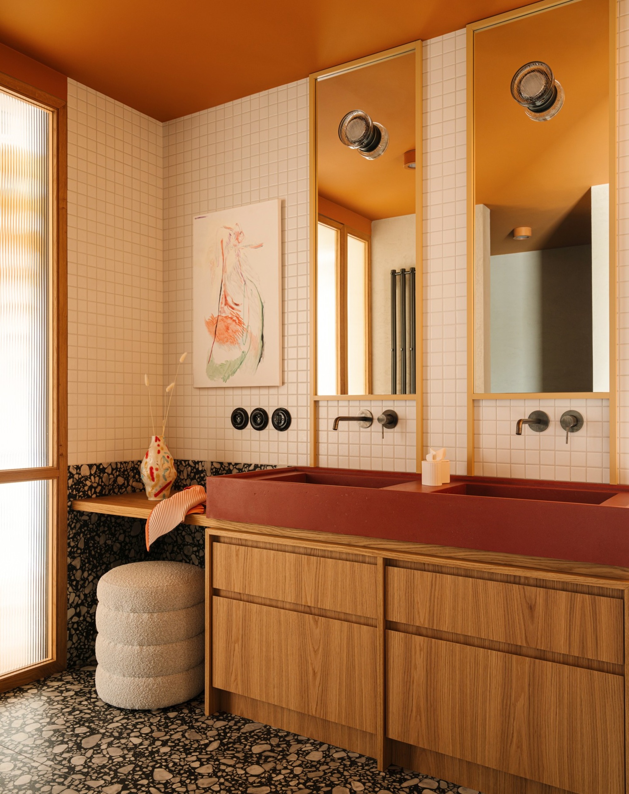 mid century modern inspired bathroom with terrazzo floors, red sink, timber vanity with storage, two wall mirrors with built-in light, chrome taps, an artwork, white square tile walls, and an ottoman vanity station