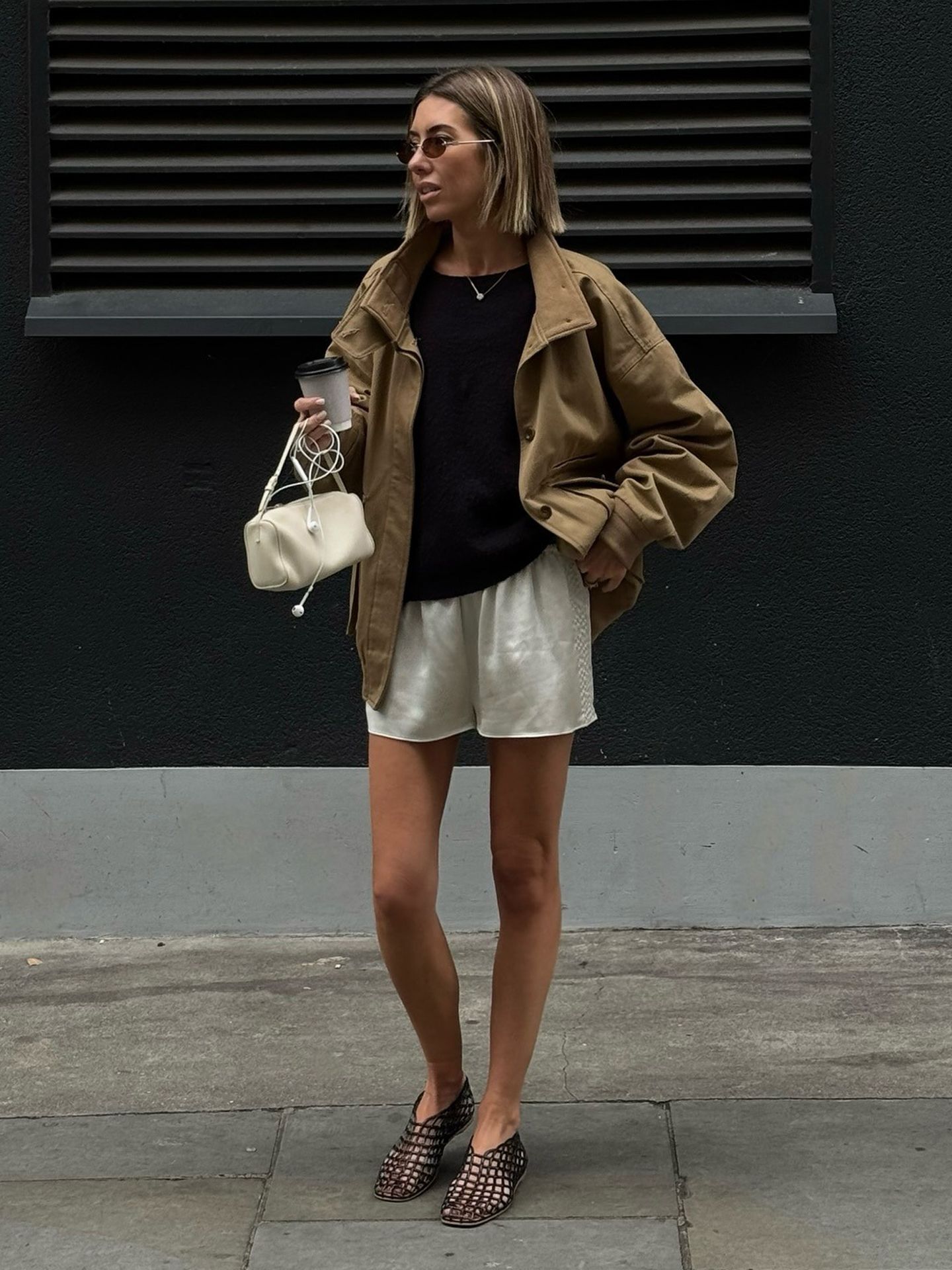 British style influencer Hannah Lewis poses in London wearing a tan jacket, black top, ivory silk shorts, mini white bag, and black jelly flats