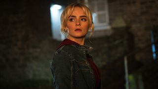 Ruby Sunday (Millie Gibson) stands in a street in semi-darkness. Her body is facing to the right of the picture, but her head is turned over her right shoulder and she's looking behind her. She's wearing an indigo denim jacket over a dark red hooded top.