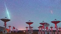 a green streak of light zooms among the stars above a set of antenna dishes in the desert