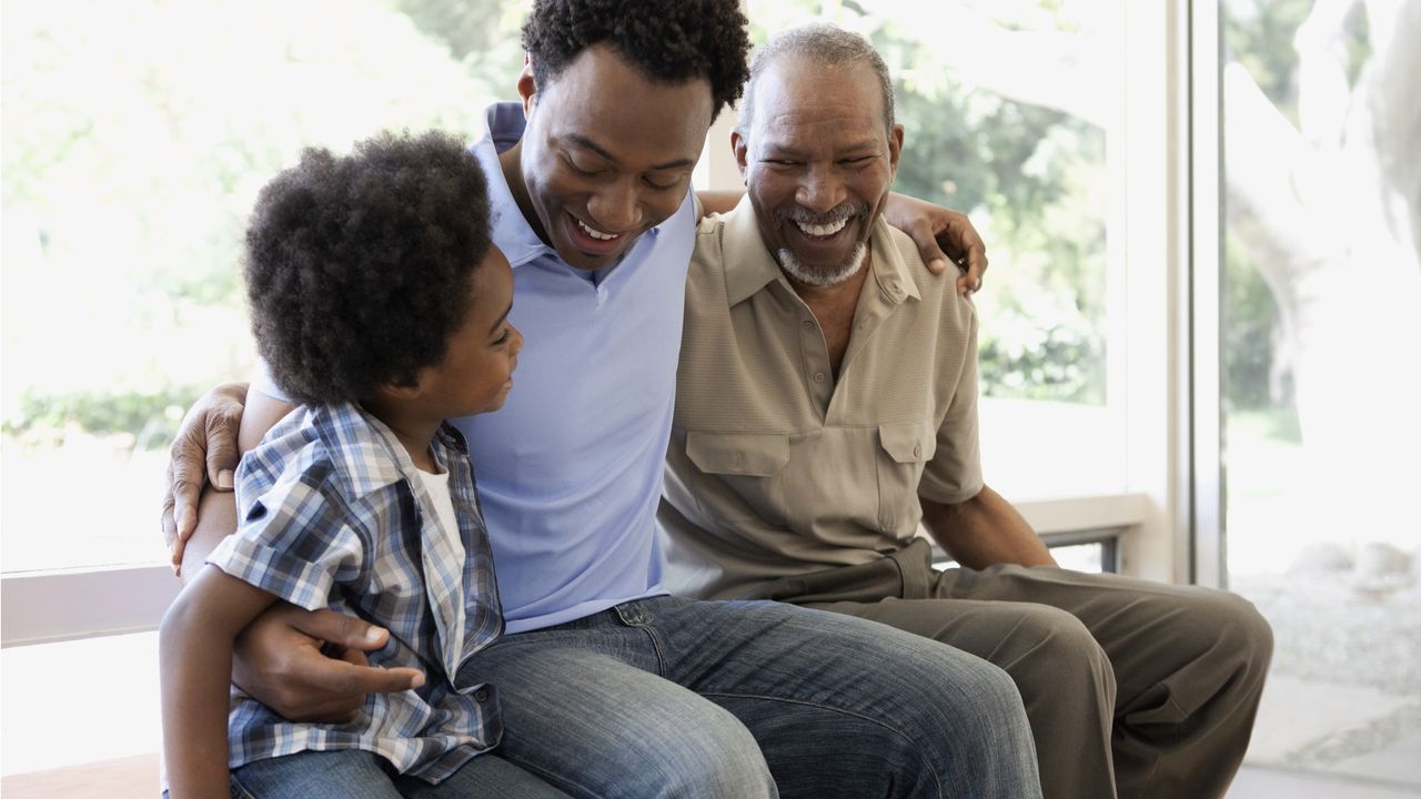 Three generations of men (grandson, son and dad) sit with their arms around each other on a bench.