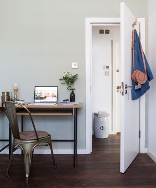 Wooden and steel desk facing wall with gold coloured chair in front of it with white open bedroom door with jean jacket and leather bag hanging on hook behind the door
