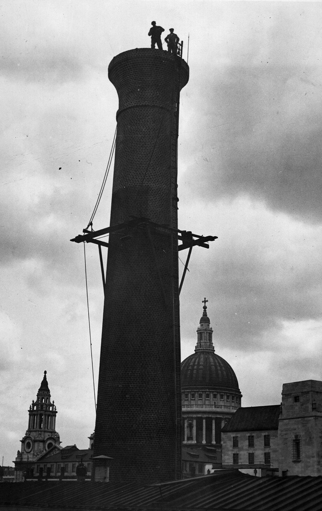 Steeplejacks are a breed apart. This pair stand atop a chimney in the City of London during a repairing operation, with St Paul&#039;s Cathedral as their backdrop.