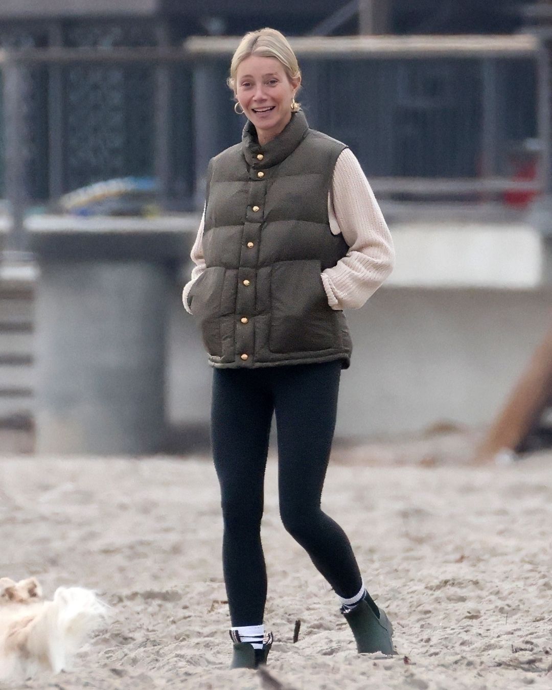 Gwyneth Paltrow walks down the beach wearing black leggings with olive green wellies and an olive green vest top and a beige jumper with gold hoop earrings.