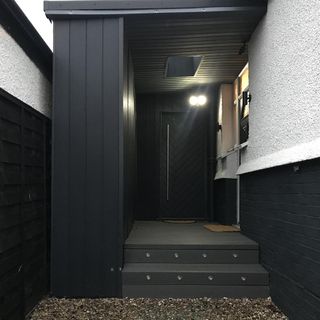 a black porch with black walls, door and stairs, with black and white exterior walls and dark gravel leading to the stairs