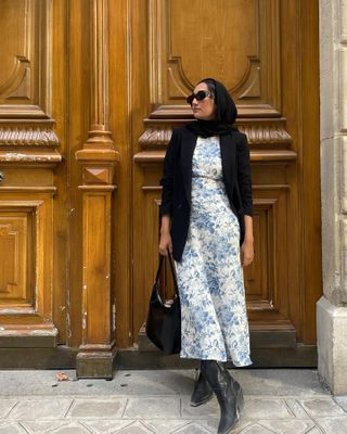Woman wears white-and-blue floral maxi sundress with black blazer, black hijab, black boots, and black sunglasses.