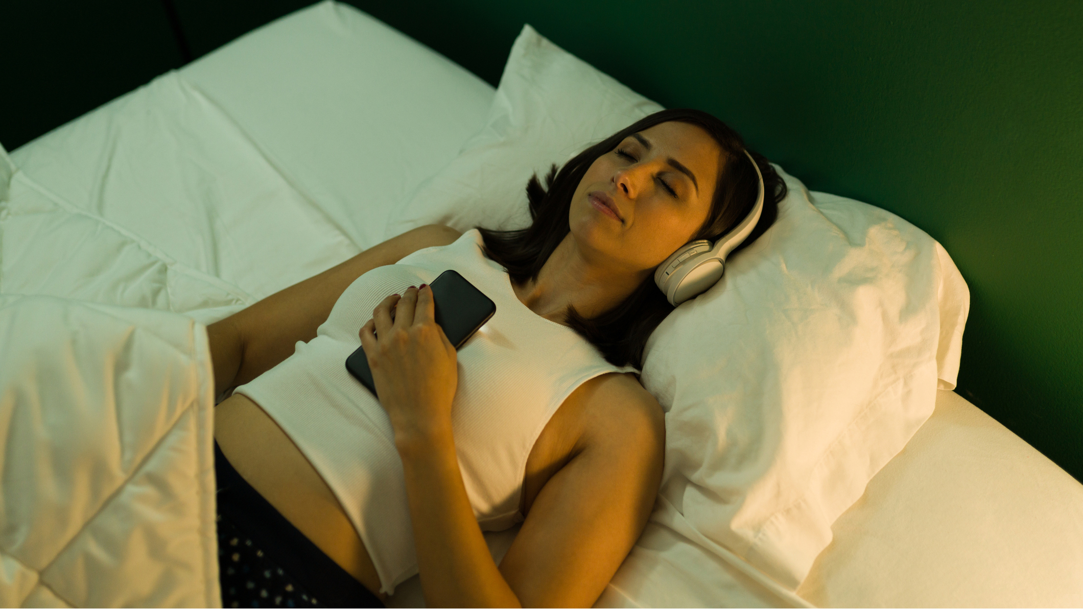 Woman lying in bed wearing over-ear headphones and holding phone on her chest