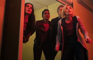 Sarah (Aisling Bea), Yaz (Mandip Gill), Dan (John Bishop) and The Doctor (Jodie Whittaker) emerge from a doorway in a storage facility, looking very alarmed by what awaits them just off-camera...