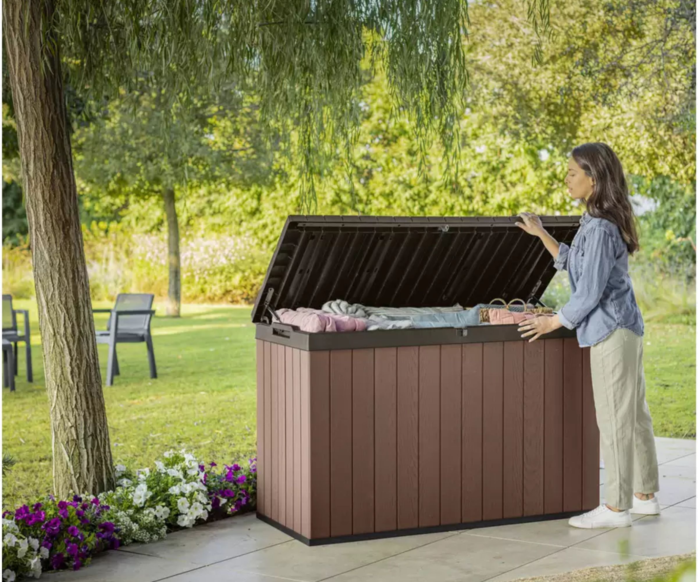 Woman opening a large brown wood-effect outdoor storage box on a sunny patio surrounded by flowers and trees.