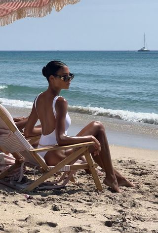 A woman wearing a low-back white one-piece swimsuit with black sunglasses while sitting on the beach.