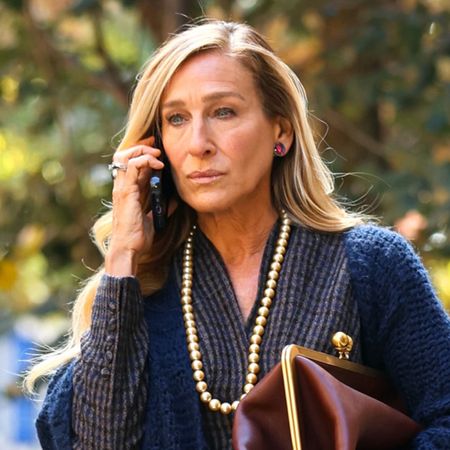 A photo of Sarah Jessica Parker on the set of And Just Like That wearing a navy shawl, patterned blouse, A-line denim skirt, and Coach's new Glovetanned Leather Kisslock Frame Bag in burgundy. 