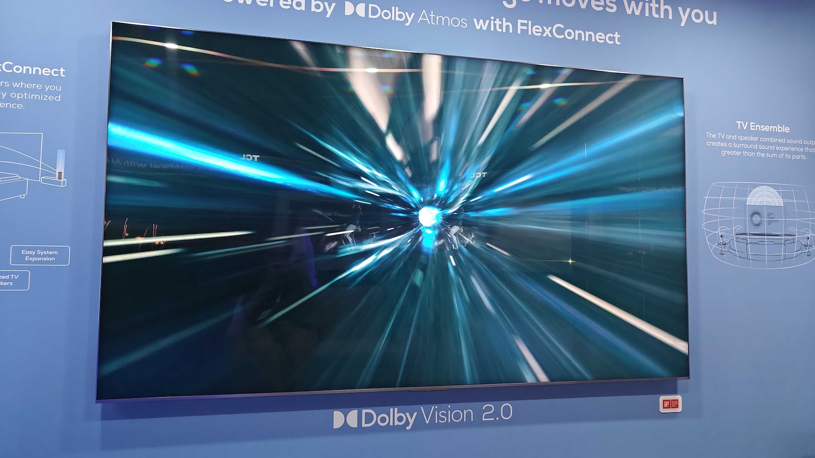 A photo of a 98-inch TV mounted to a blue wall at a trade show. Various logos and text surround the screen, including 'Dolby Vision 2.0'