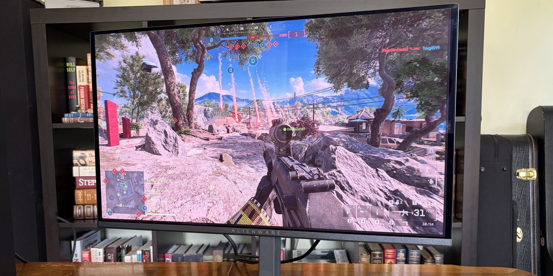 A gaming monitor shows a character aiming a rifle in a vibrant outdoor scene with rocky terrain and trees, under a blue sky.