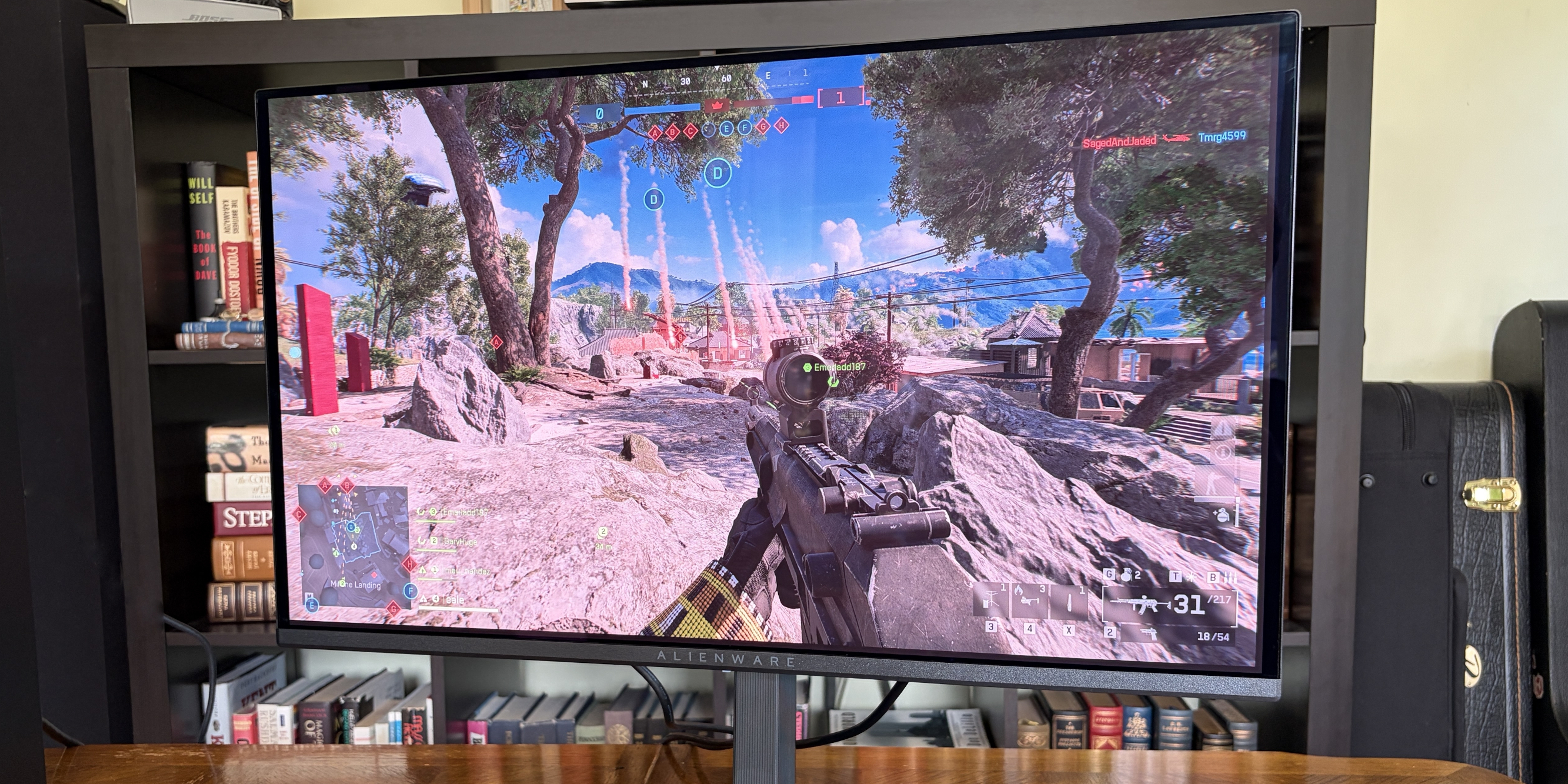 A gaming monitor shows a character aiming a rifle in a vibrant outdoor scene with rocky terrain and trees, under a blue sky.