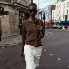 Model wearing brown cardigan and white trousers, walking down street.