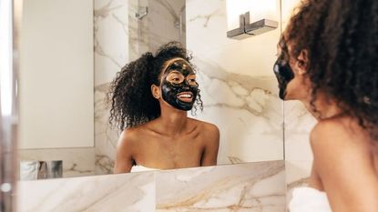 A woman wearing a clay mask smiles at her reflection in the mirror.