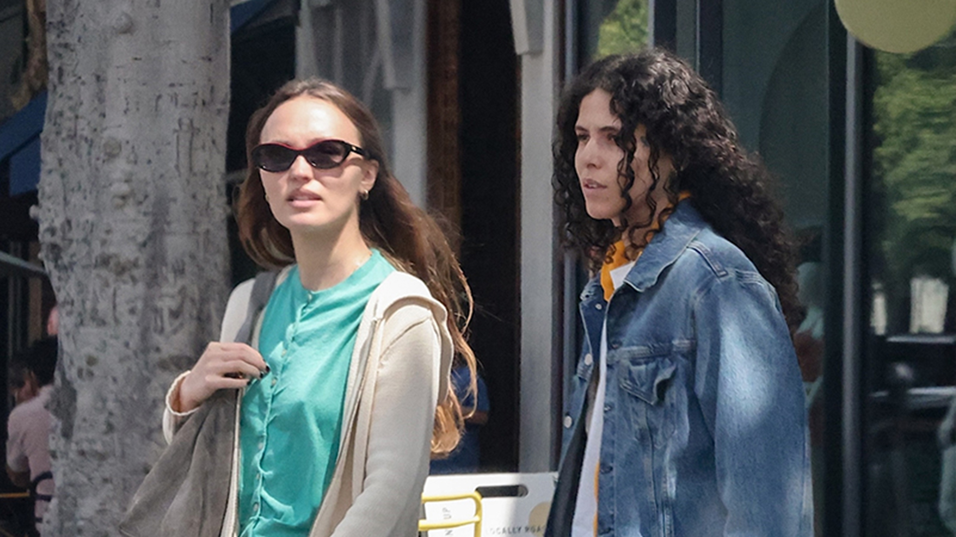 Lily Rose Depp wearing a teal shirt, beige hoodie, indigo jeans, suede shoulder bag, and tan suede loafers.