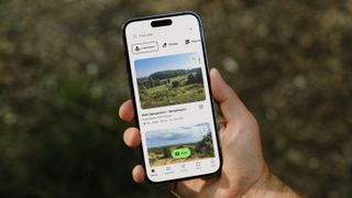 A hiker's hand holding a phone showing routes from AllTrails on the screen