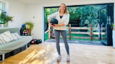 Fitness writer Maddy Biddulph stands in her living room holding a medicine ball and smiling at the camera.
