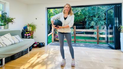 Fitness writer Maddy Biddulph stands in her living room holding a medicine ball and smiling at the camera.