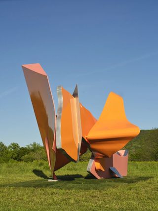 In a sunny park, an abstract, large-scale installation seemingly cut out of some sort of metal and covered in orange paint adds a fantastical feel to the outdoor location.