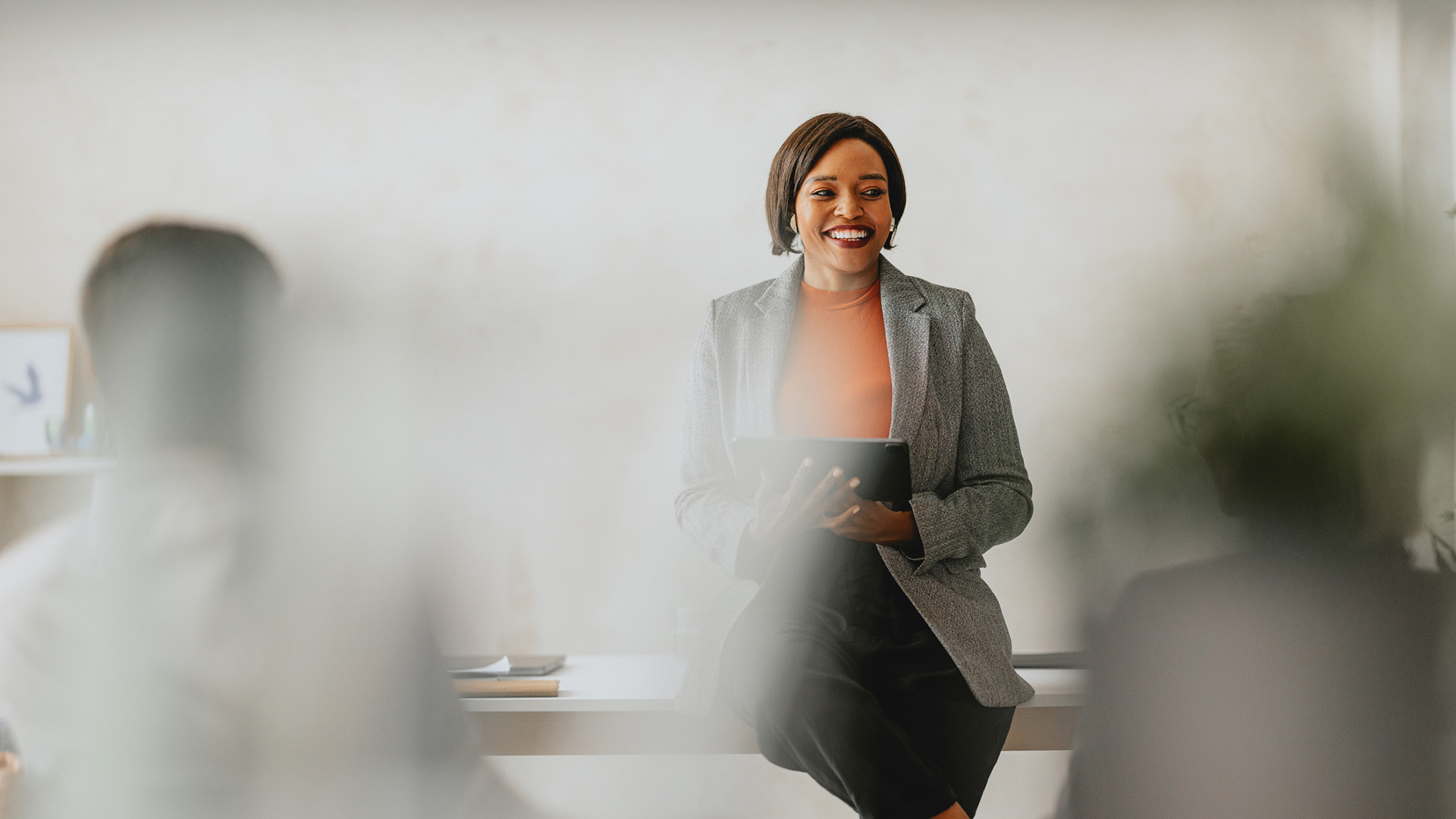 A female executive sat on a table in front of colleagues, smiling holding a tablet and wearing a blazer over an orange jumper and holding a tablet.