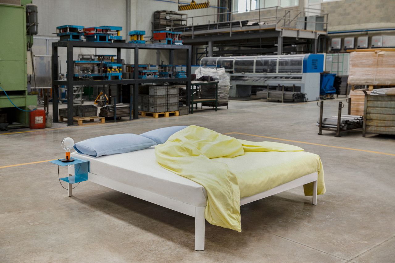 A white bed with light blue pillows and yellow sheets, including a built in side table in blue. The bed is shown inside a factory space