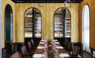 Dining room with yellow wall tiles around arched windows of wine storage area, long table and rattan chairs