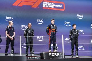F1 drivers Lewis Hamilton, Max Verstappen, and George Russell stood on podiums in front of a wall bearing multiple iterations of the AWS logo. An orange LED sign above the drivers identifies the race as the Formula 1 Gran Premio de España 2023.