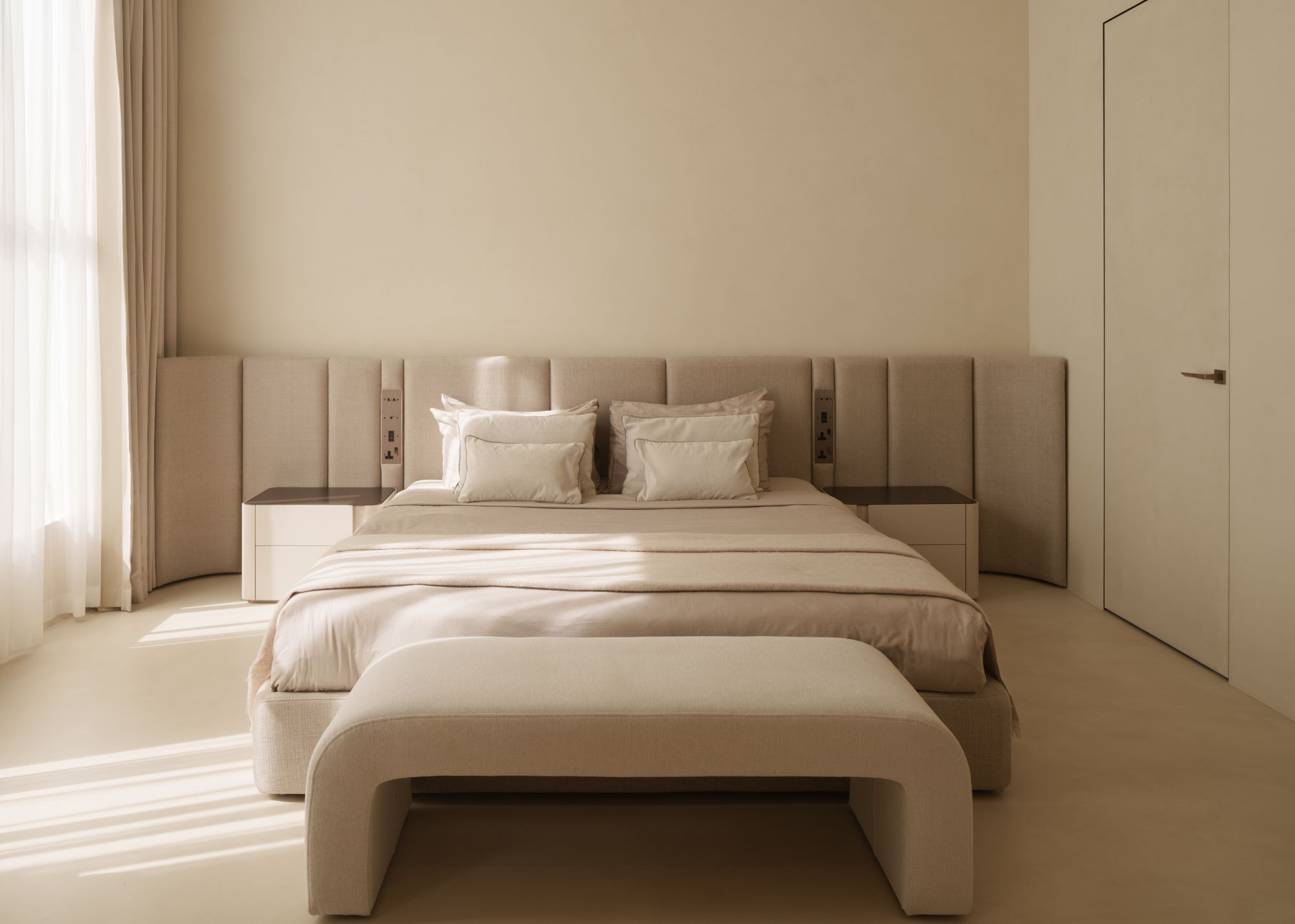A neutral bedroom with a custom built-in headboard along the entire back wall with integrated plug sockets and a cream bench sits at the foot of the bed