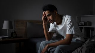 A man sits on the side of his bed in a dark room with his head in his hands because he's experiencing distress from being unable to sleep