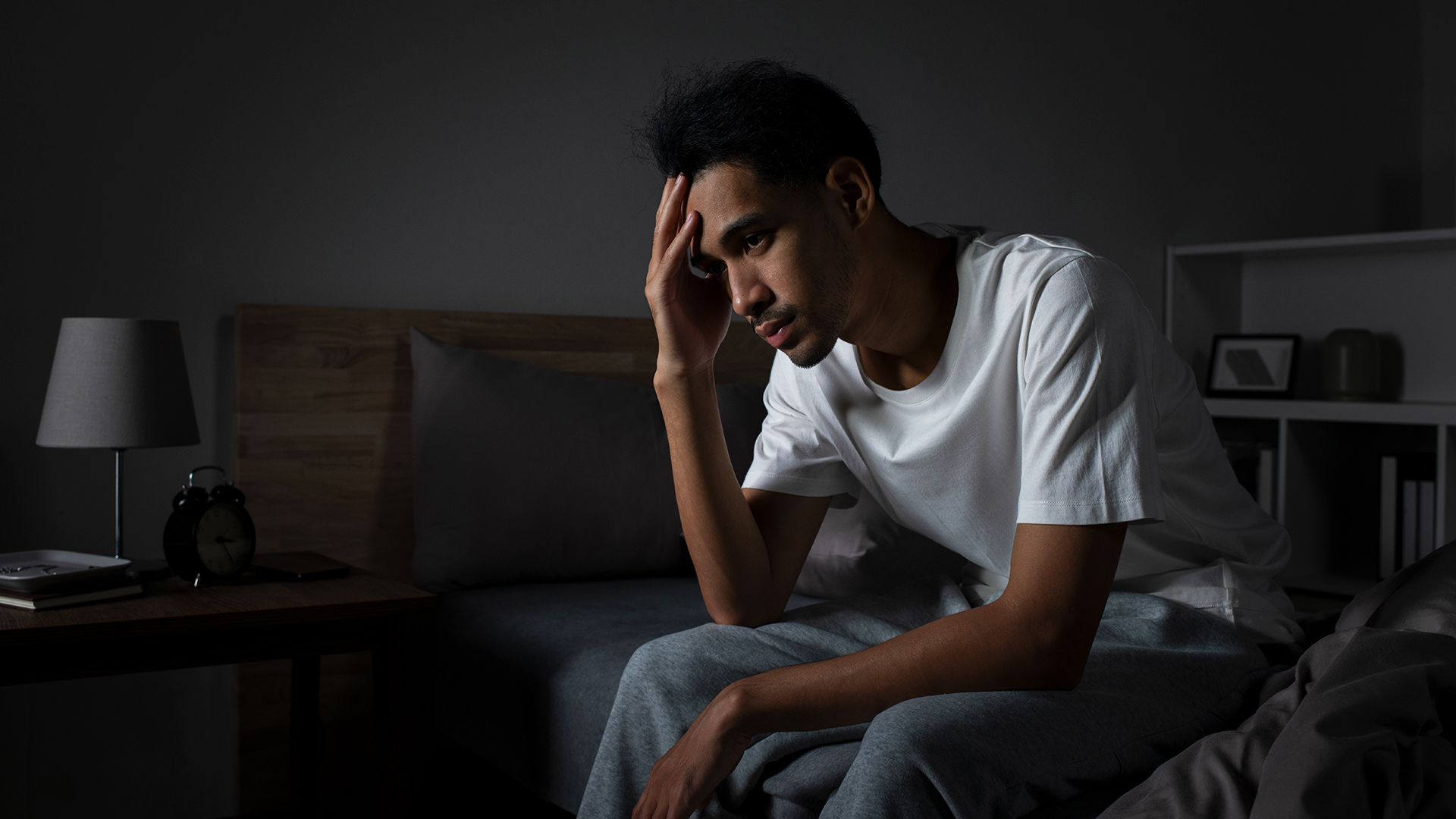 A man sits on the side of his bed in a dark room with his head in his hands because he&#039;s experiencing distress from being unable to sleep