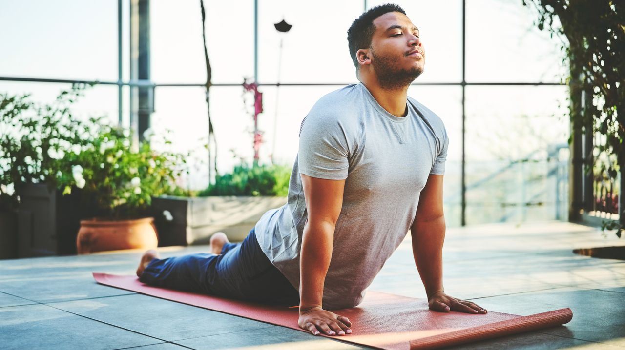 A man in a t-shirt and leggings performs upward dog on a yoga mat in front of a glass-panelled wall. He is lying down on the mat, with his legs behind him, hands on the floor and his arms straight, so that his torso is held upwards. His eyes are closed and his face is bright with sunlight. Behind him we see windows, glass panels and leafy potted plants.