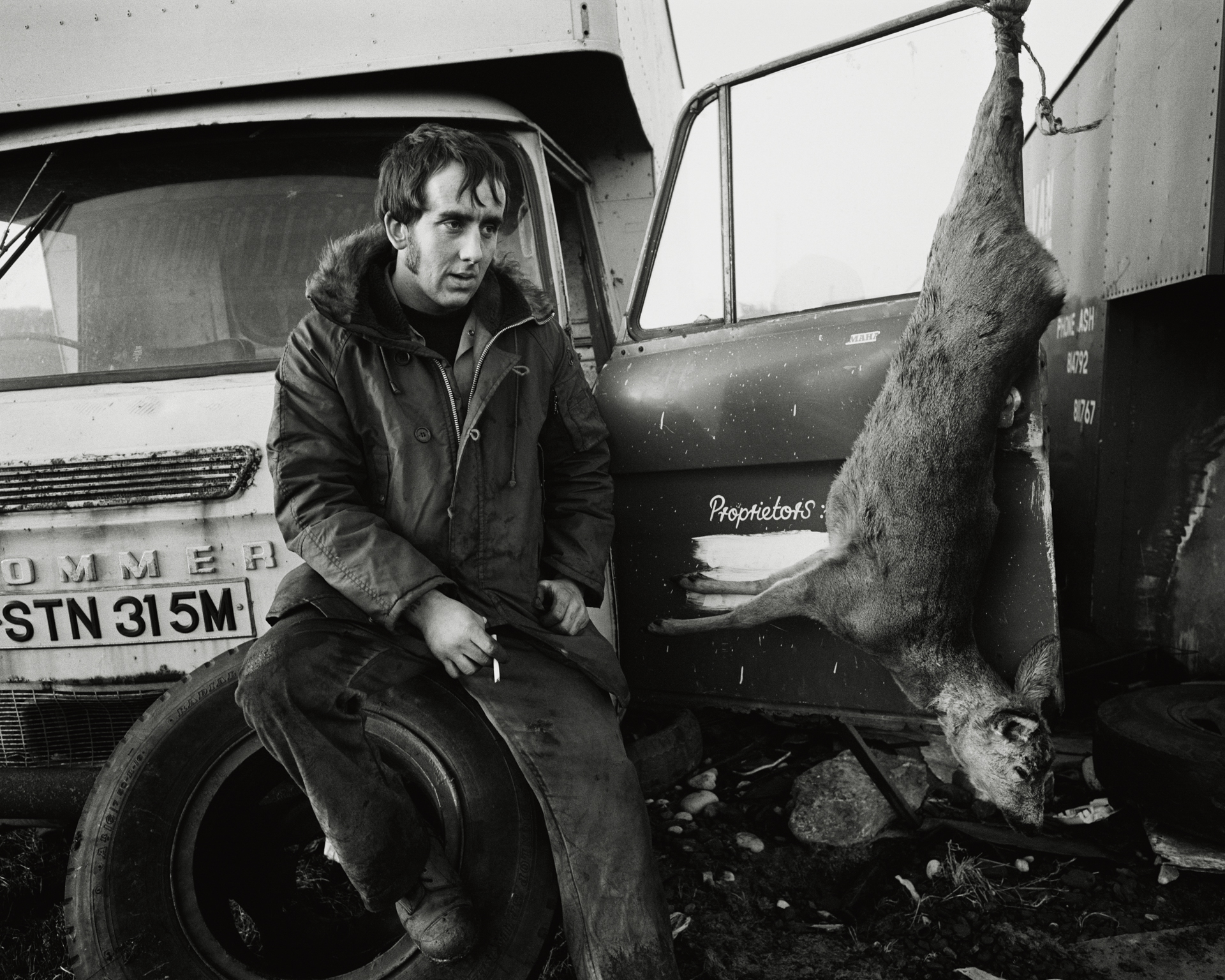 Black and white photograph of a young man sitting on a tyre beside an old lorry, a dead deer hanging from the vehicle beside him, taken at the seacoal camp, Lynemouth, Northumberland, in 1984.