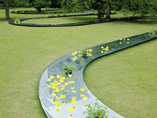 A park area with a winding glass piece of art work that runs along the grass with flowers on top.